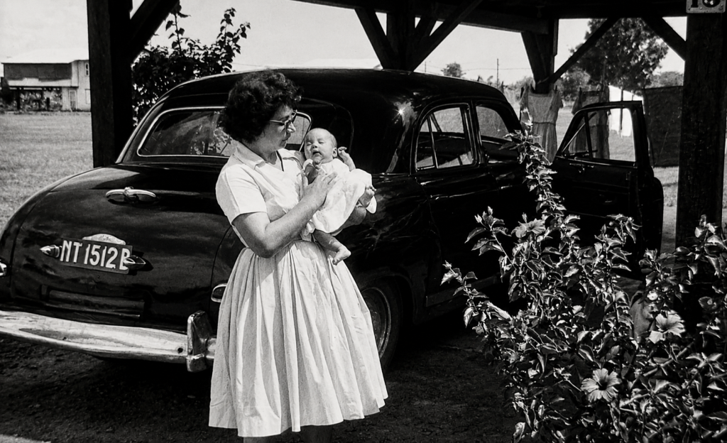 Black-and-white photograph of Ramona as a baby being held by her mother beside the family car.